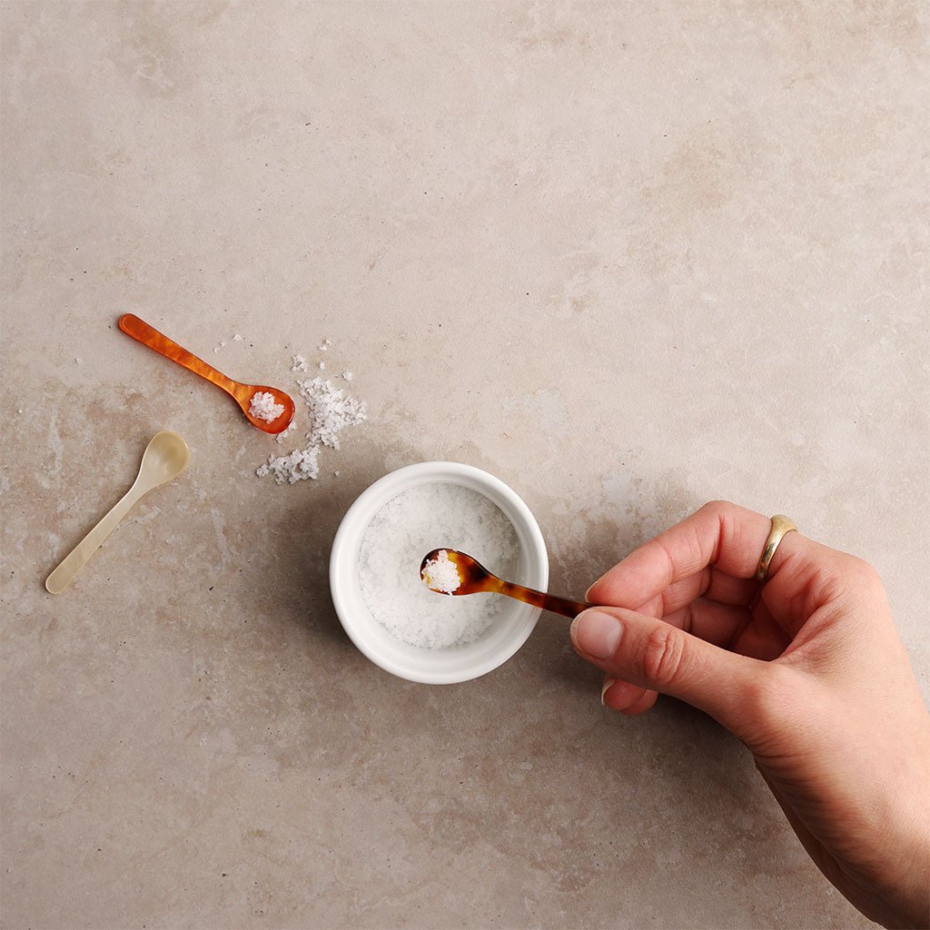 Hand holding a small acrylic spoon with salt over a small white bowl on a beige surface.