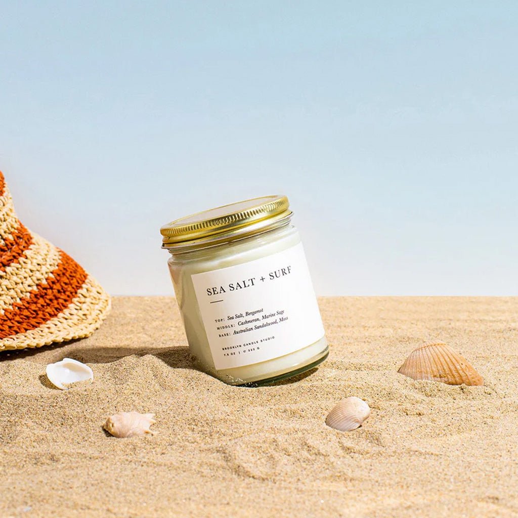 Candle labeled 'Sea Salt + Surf' on sand with shells and a hat in the background