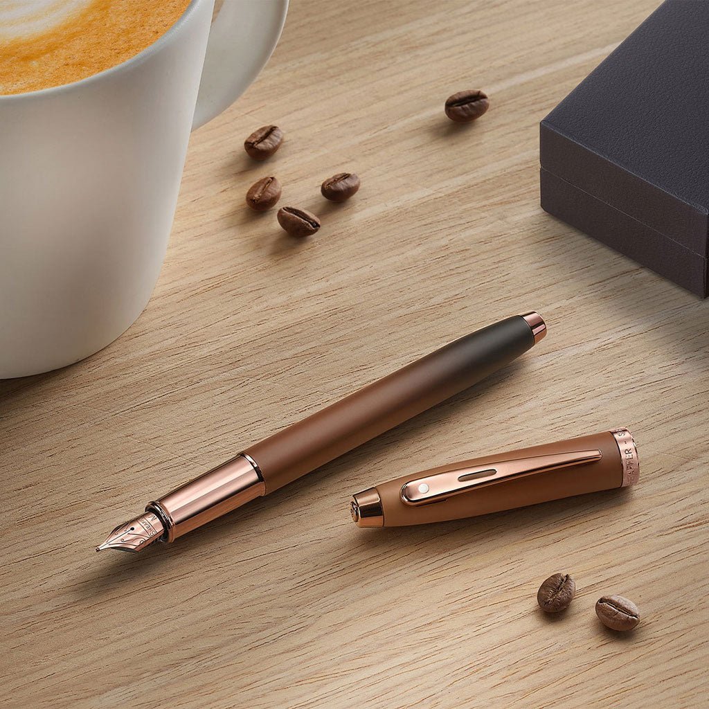 Brown rose gold pen on a wooden surface with coffee beans and a cup of coffee.
