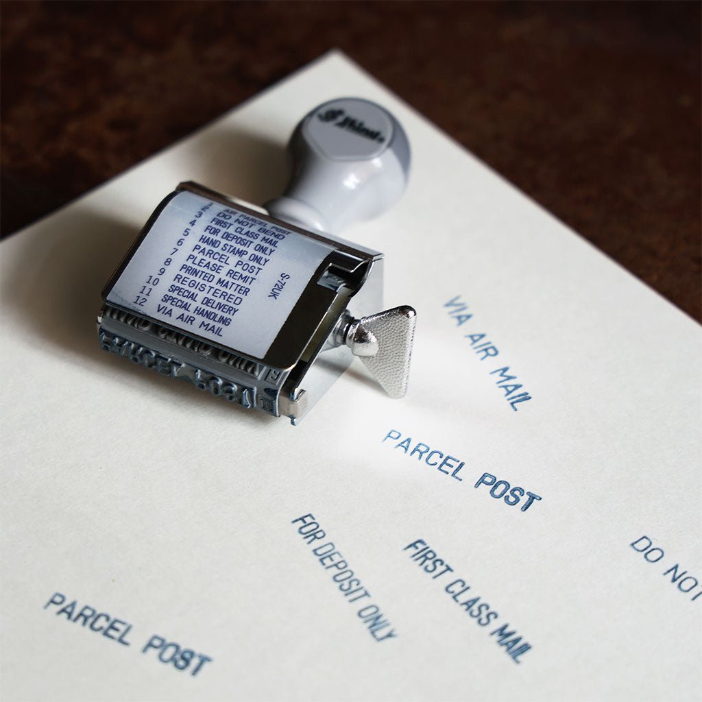 A rotating, handheld stamp lying on a piece of paper that has been stamped with phrases like PARCEL POST, VIA AIRMAIL, and FIRST CLASS MAIL.