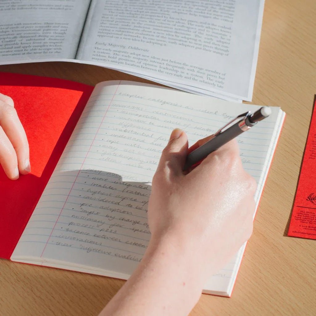 Hand holding pencil writing in red exercise notebook with lined pages on wooden background