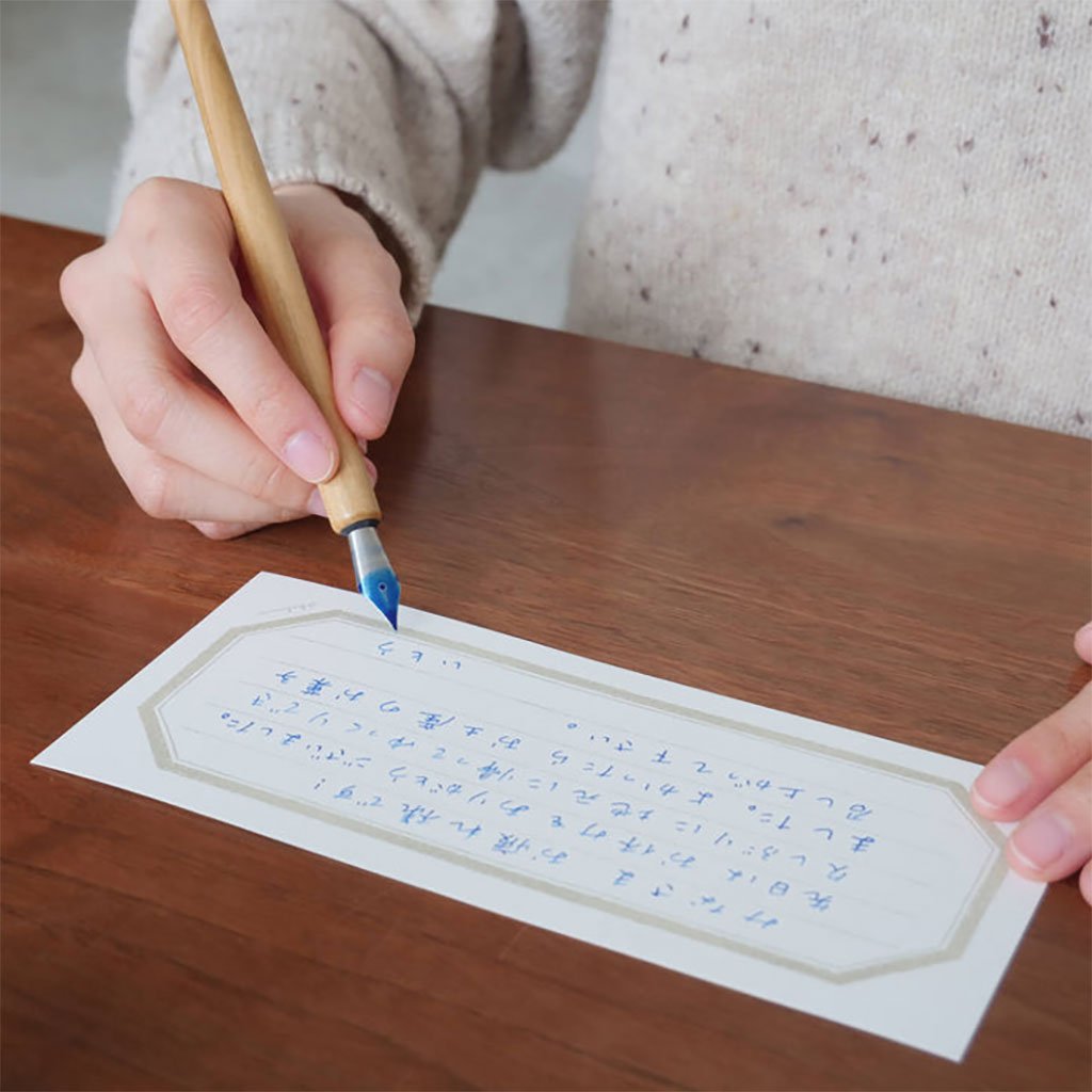 Person writing on a piece of paper with a dip pen on a wooden table.