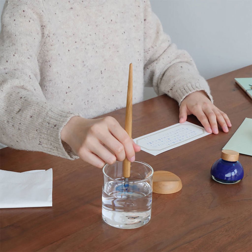 Person dipping a pen into a glass of water on a wooden table with calligraphy supplies.