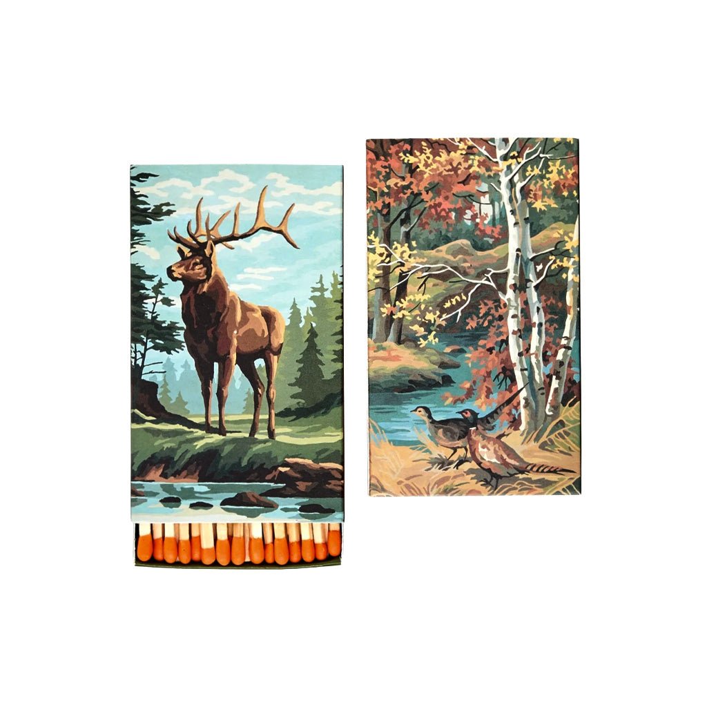 Two rectangular decorative matchboxes with woodland illustrations, shown side by side on a plain white background. The left matchbox features a brown elk standing near water, and the right matchbox shows autumn trees and two pheasants.