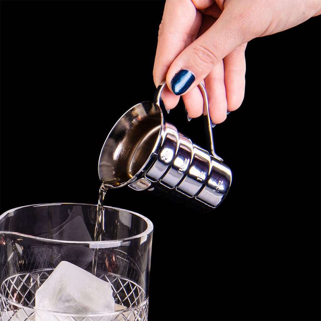 Hand pouring liquid from a metal jigger into a glass with ice on a black background