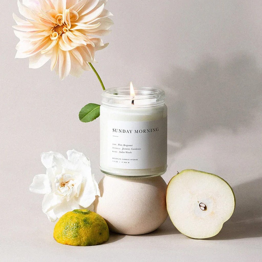 Candle labeled 'Sunday Morning' with flowers and a fruit on a light background