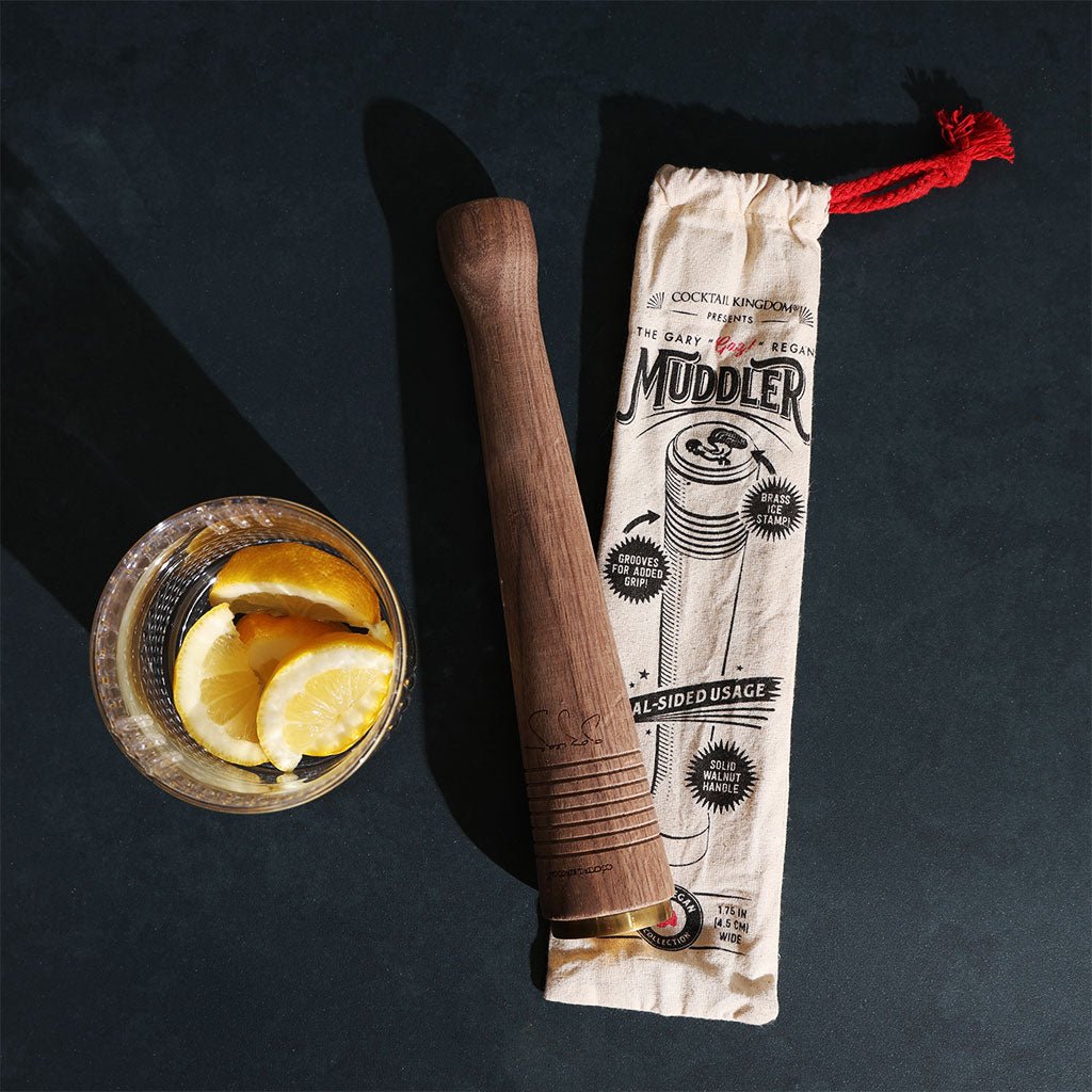 Wooden muddler next to a bag labeled 'Muddler' on a dark surface with a glass of sliced lemons.