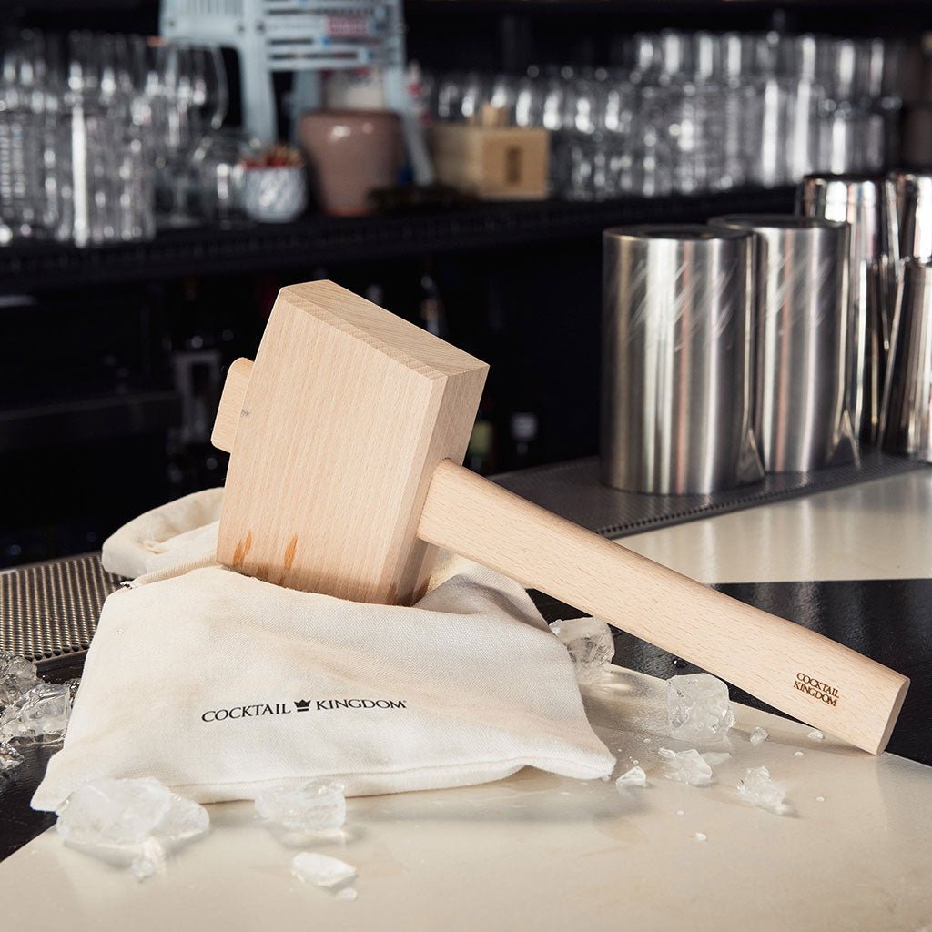 Wooden muddler on a white cloth with 'Cocktail Kingdom' branding, set against a bar background.