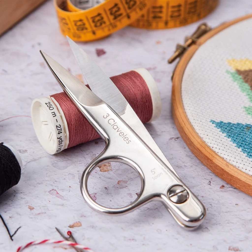 Scissors with '3 Claveles' branding on a table with spools of thread and a cross-stitch hoop.