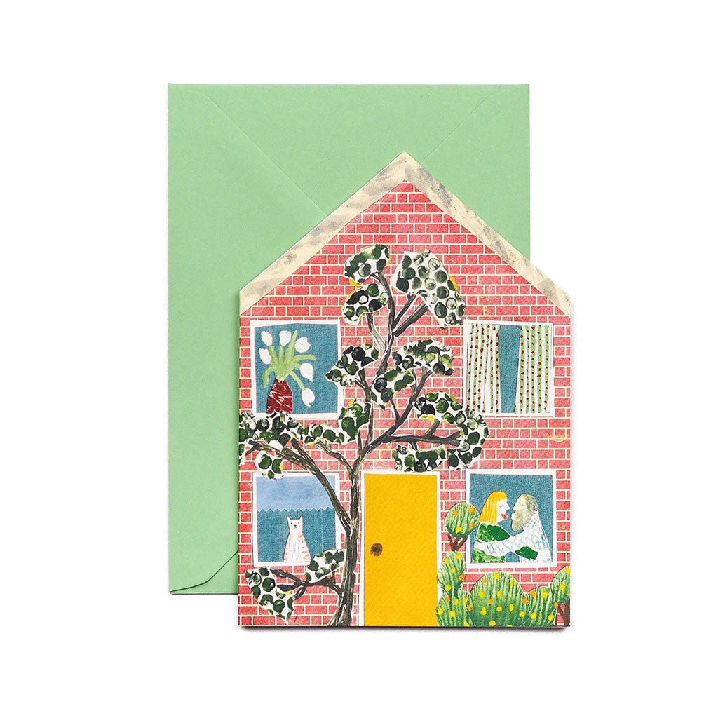 Greeting card with a colorful illustration of a house and tree, with people, a cat, and flowers in the windows, shown with a green envelope.