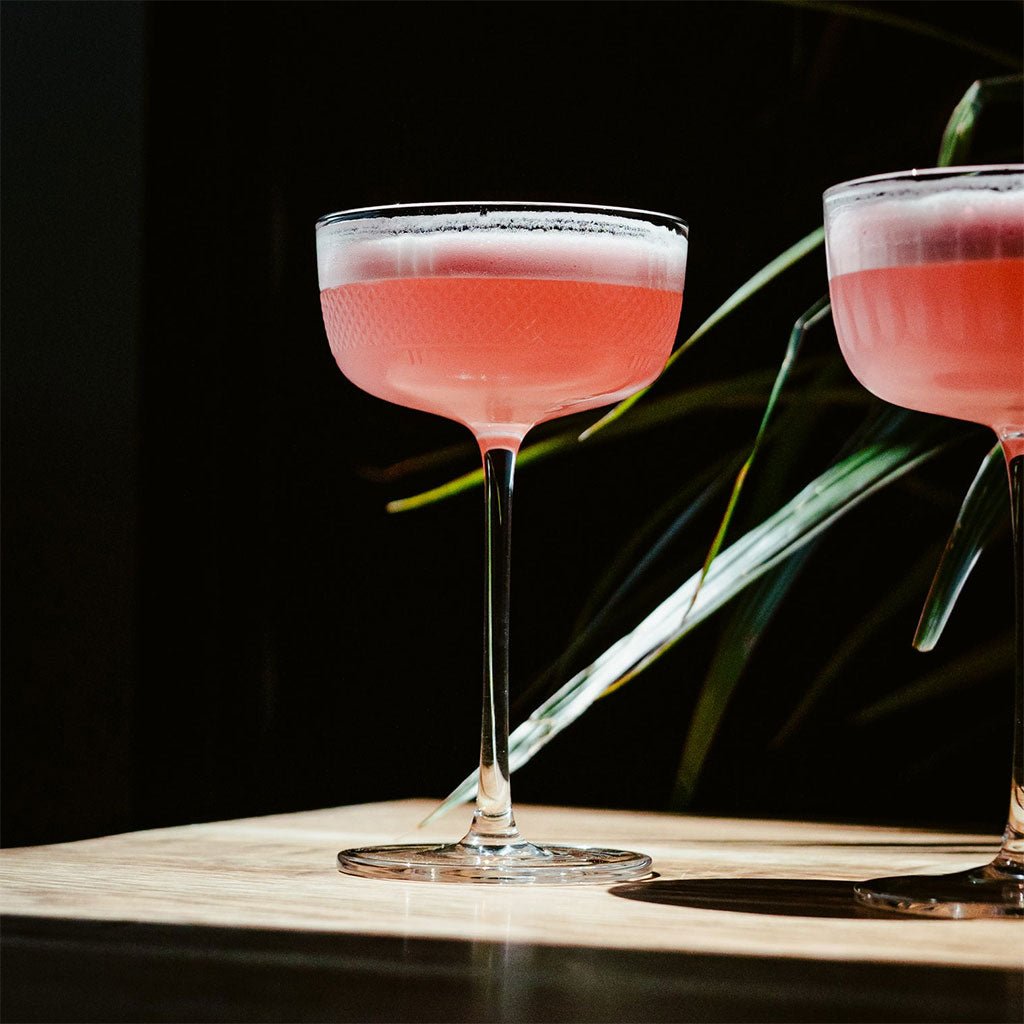 Two pink cocktails in stemmed coupes on a dark background with green leaves.