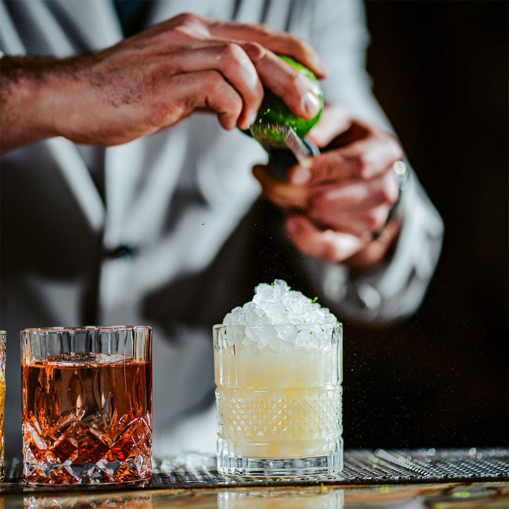 Person preparing a cocktail with ice and a lime, with another drink in the foreground.