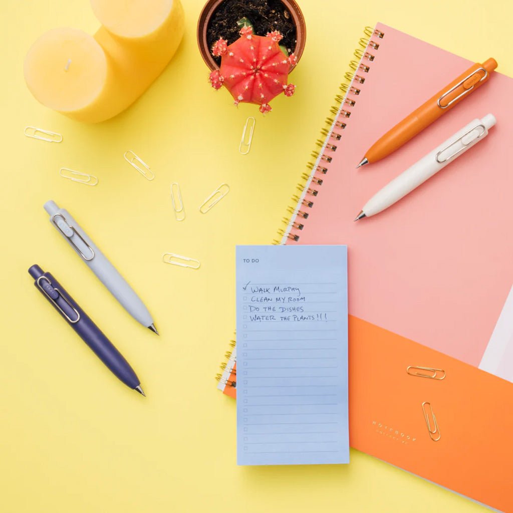 Four uniball one p gel pens in various colors next to office supplies on colorful desk background