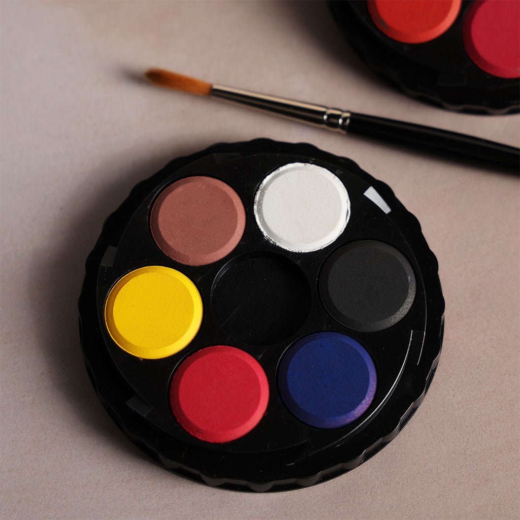 A close-up of a round plastic tray with six watercolor paints in it, with a paint brush nearby for scale.