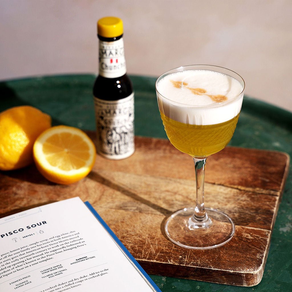 A pisco sour cocktail with a thick foam head, in a Nick and Nora glass surrounded by cocktail making accessories.
