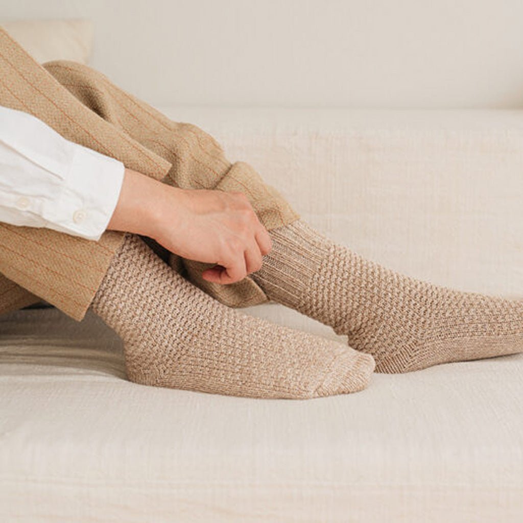 Hand pulling up beige knitted socks onto legs on light fabric couch