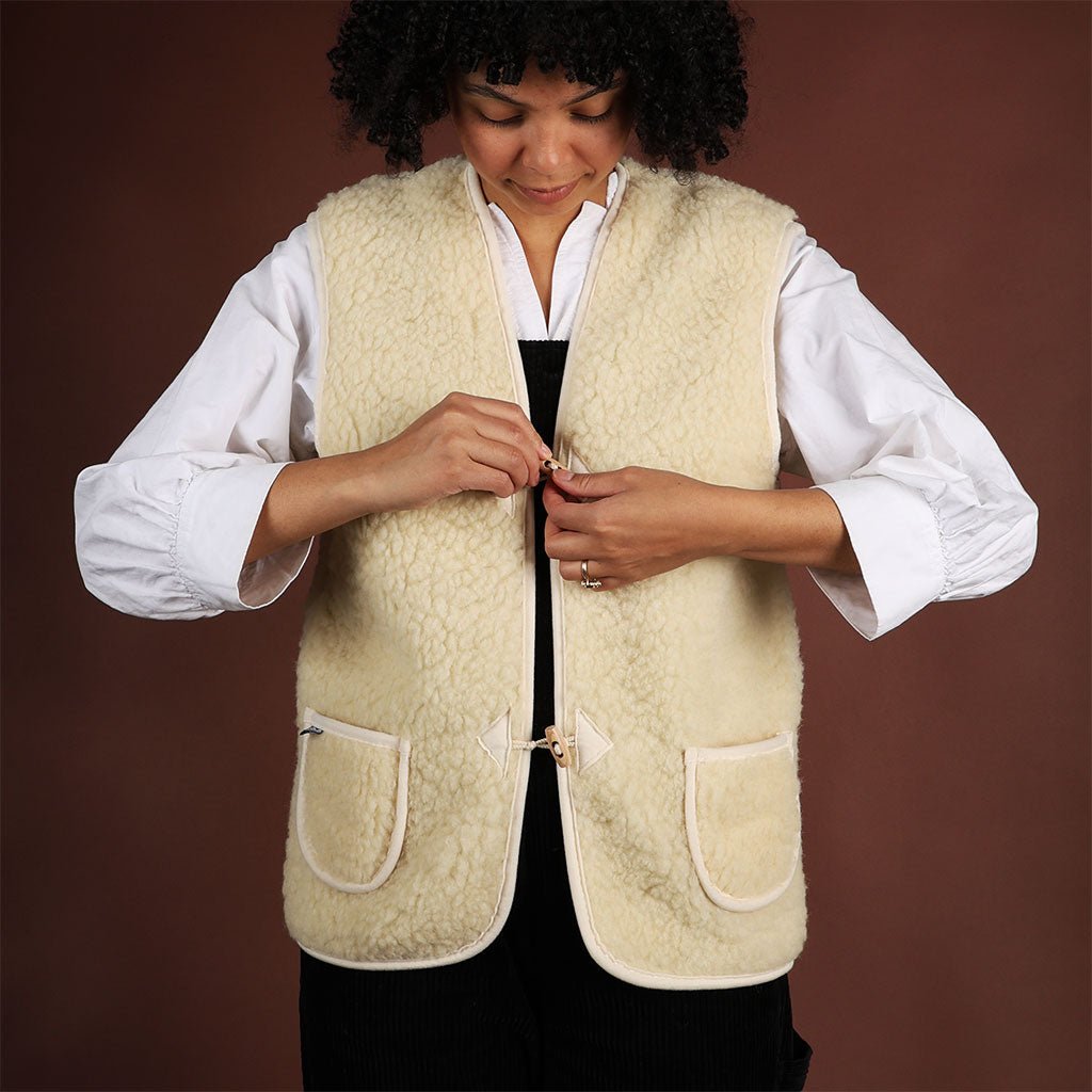 A model wearing a natural woolen vest over a white shirt and black jumper. She is fastening the toggle closure.