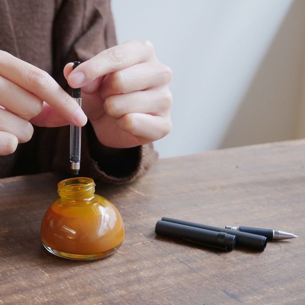 Person filling a pen with orange ink from a small bottle on a wooden surface.