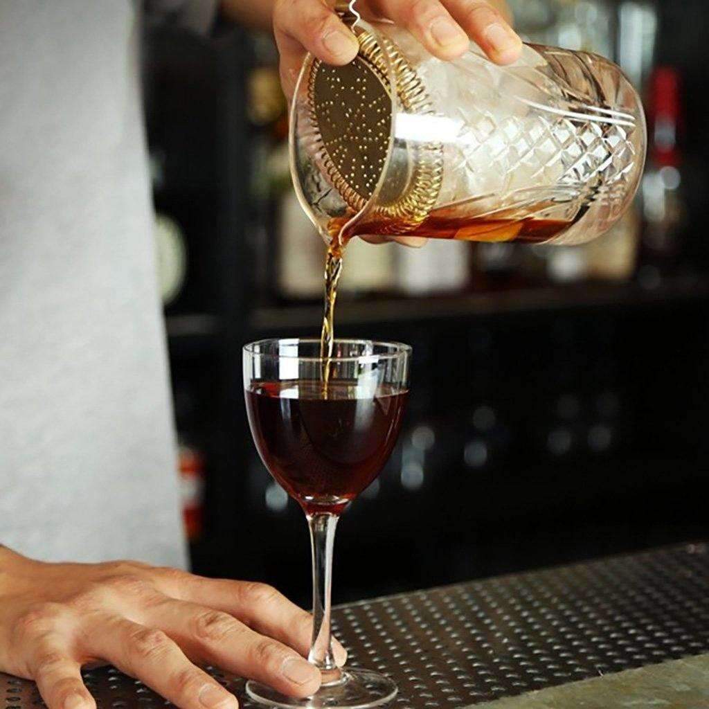 Hand pouring drink through hawthorne strainer into glass on bar top