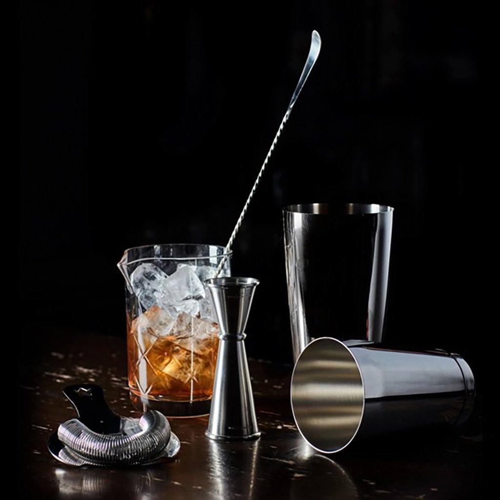 Bartending tools including a shaker, jigger, and mixing glass with ice on a dark background