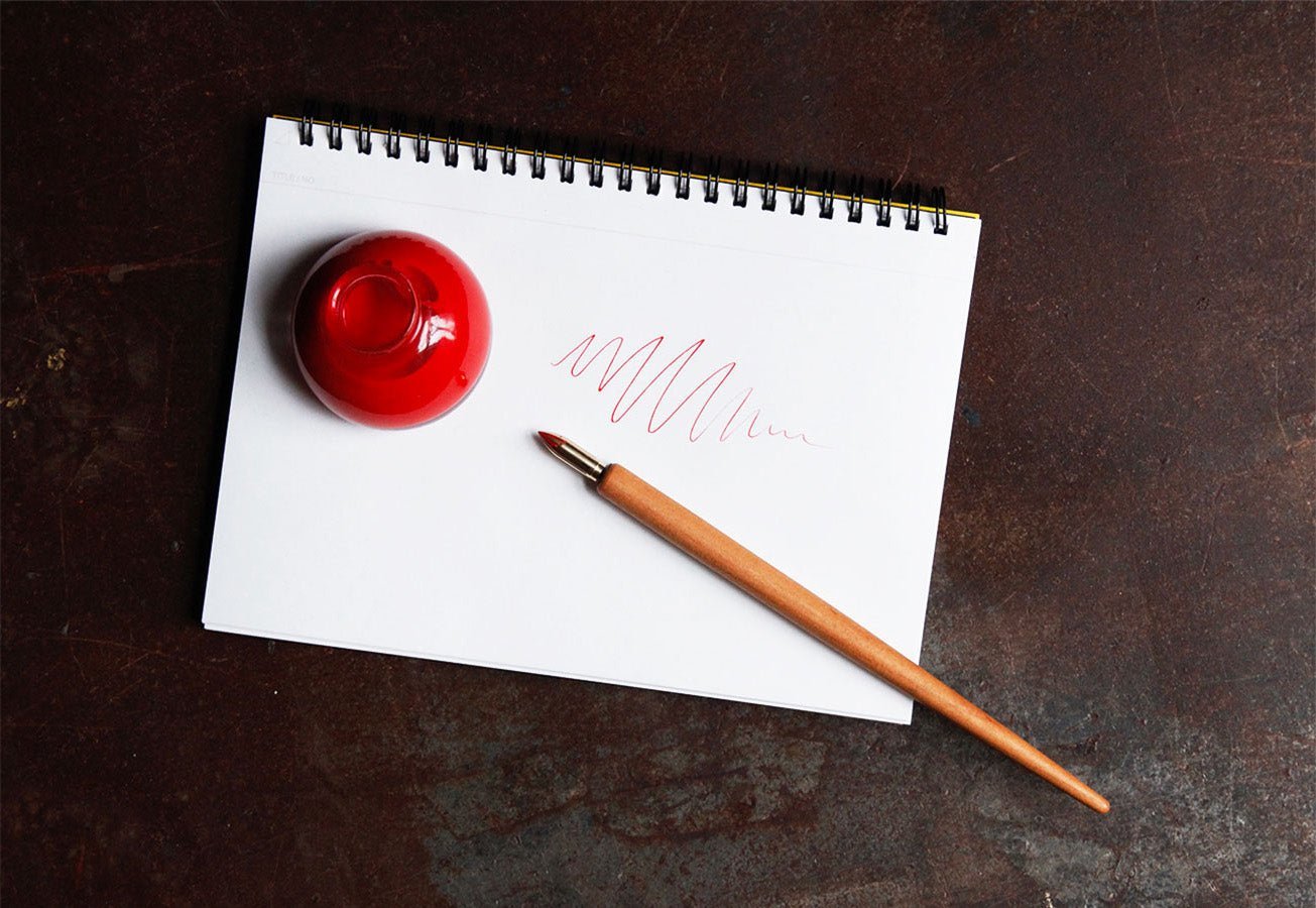 Kakimori wooden nib holder next to bottle of red ink on pad of paper on dark background