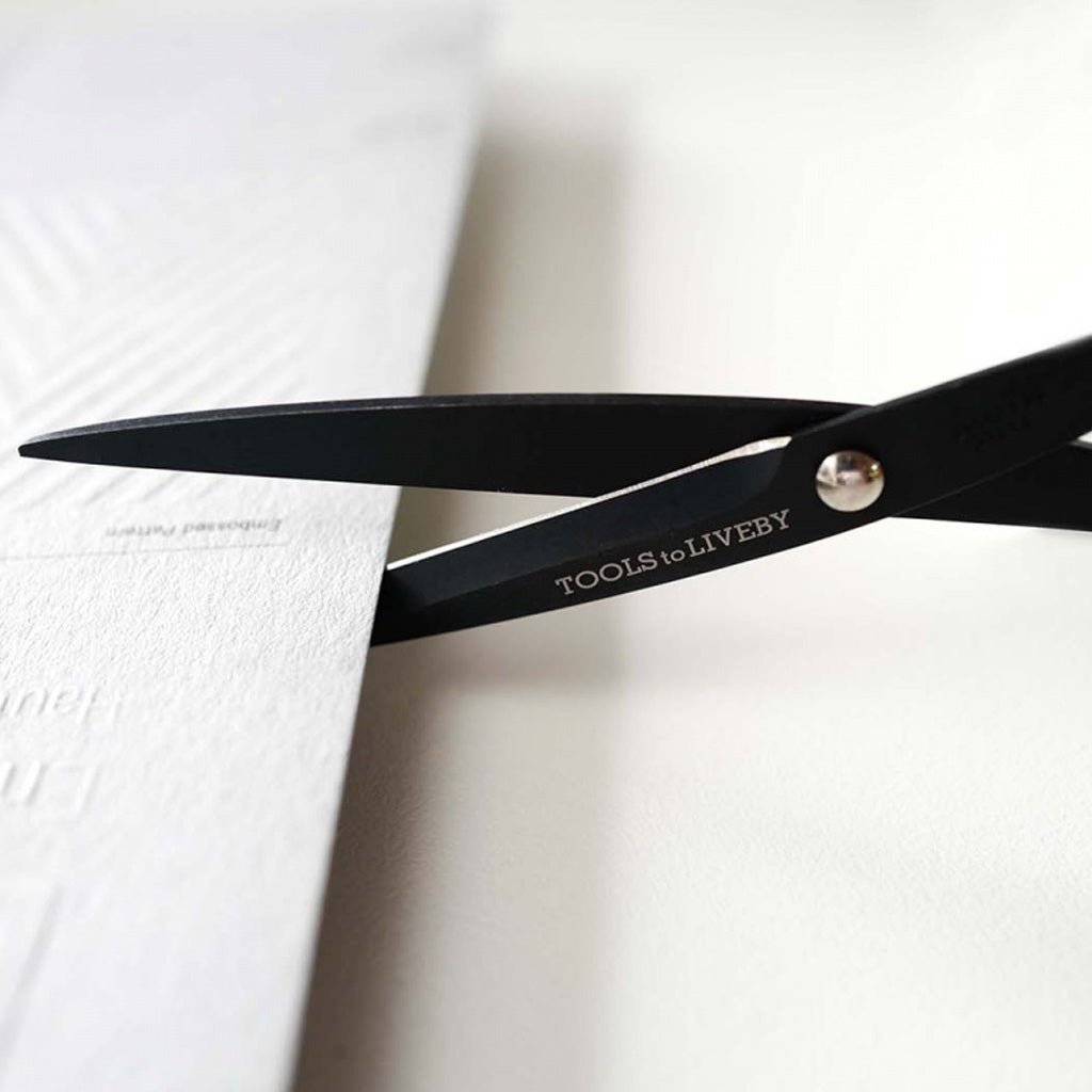 Black scissors with the brand name 'TOOLS TO LIVE BY' engraved on the blade, shown cutting a textured white sheet of paper.