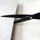 Black scissors with the brand name 'TOOLS TO LIVE BY' engraved on the blade, shown cutting a textured white sheet of paper.