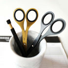 A pair of gold and black scissors and a pair of black scissors with silver rivets, standing in a white marble pencil cup alongside a black pencil on a light background.