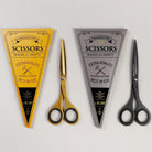 Gold-colored and matte black stainless steel scissors next to triangular packaging on gray background