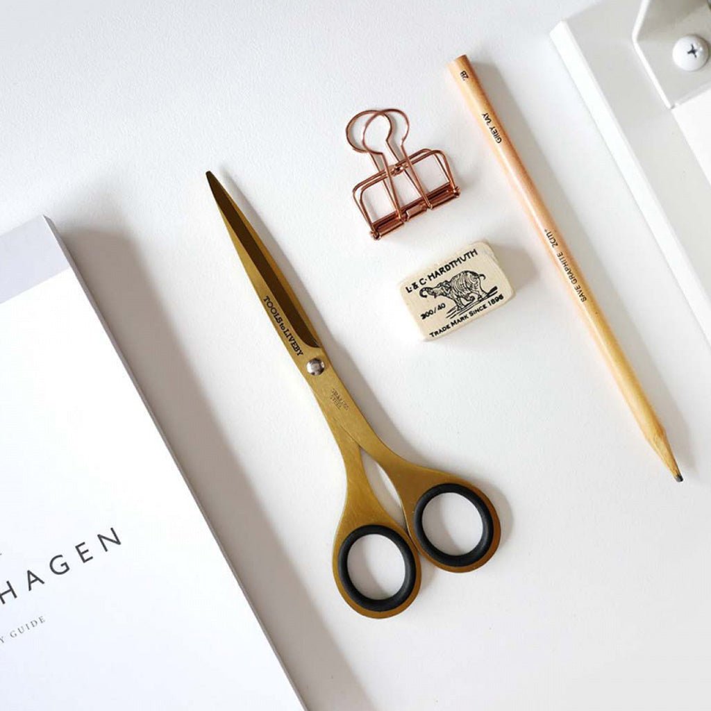 Gold-colored scissors with black-rimmed handles arranged on a white surface alongside a wooden pencil, a copper binder clip, a small eraser, and part of a white notebook.