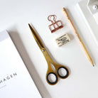 Gold-colored scissors with black-rimmed handles arranged on a white surface alongside a wooden pencil, a copper binder clip, a small eraser, and part of a white notebook.