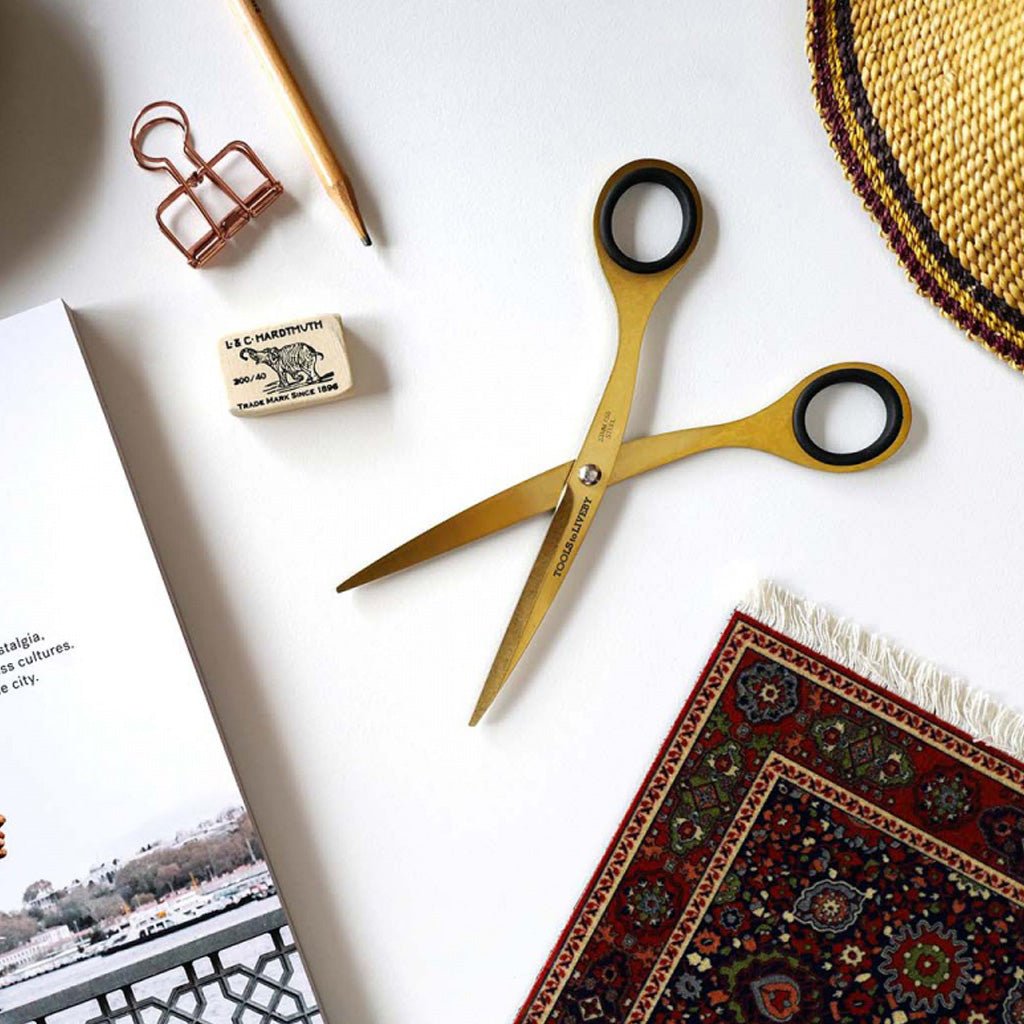 Gold scissors with black-rimmed handles placed on a white surface, surrounded by a pencil, copper binder clip, rubber stamp, book page, and patterned rug.