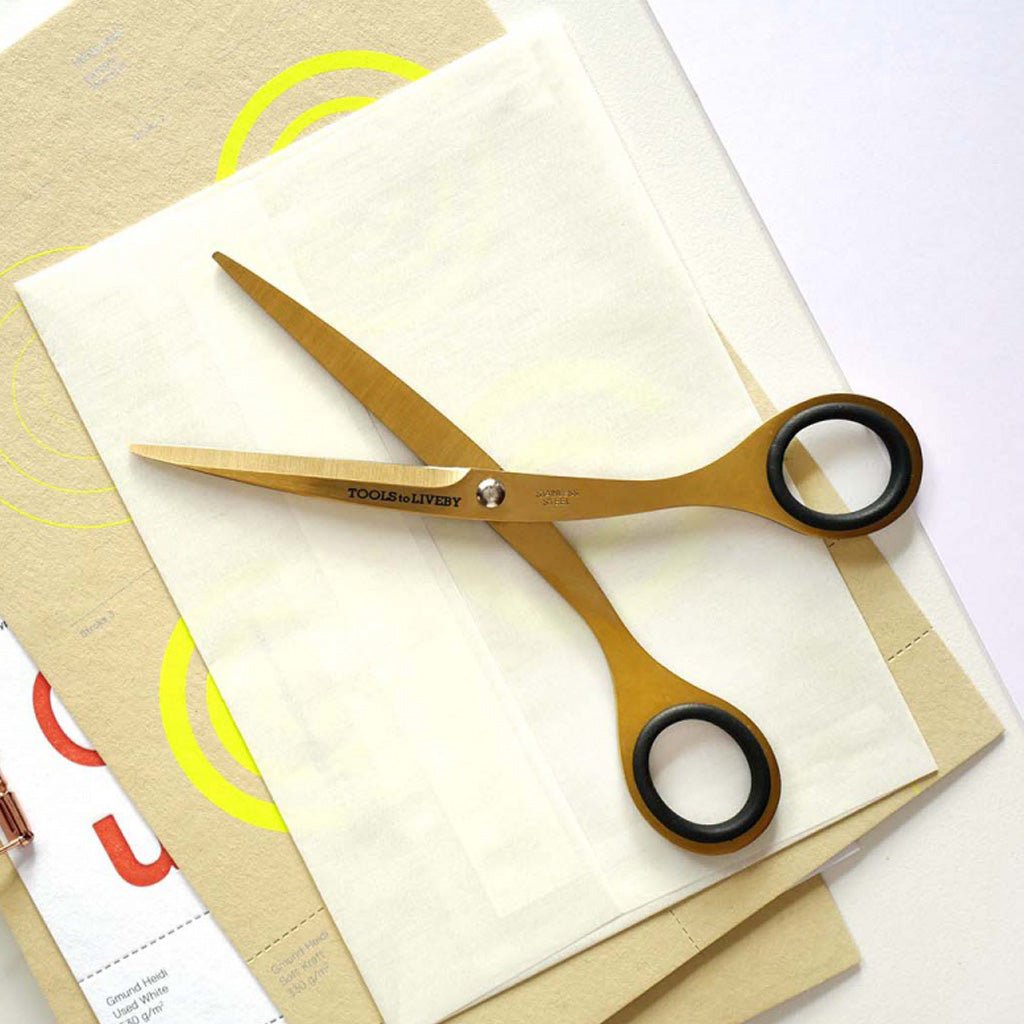 Gold-toned scissors with black ring handles resting on stacked cream-colored papers and a tan sheet with yellow circular graphics.