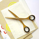 Gold-toned scissors with black ring handles resting on stacked cream-colored papers and a tan sheet with yellow circular graphics.