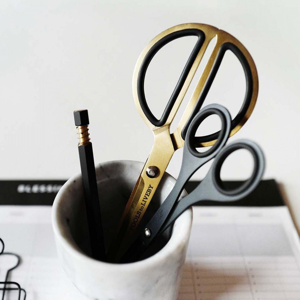 A pair of gold and black scissors with a second black pair of scissors and a black pencil resting in a small white marble cup, set against a light background.