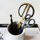 A pair of gold and black scissors with a second black pair of scissors and a black pencil resting in a small white marble cup, set against a light background.