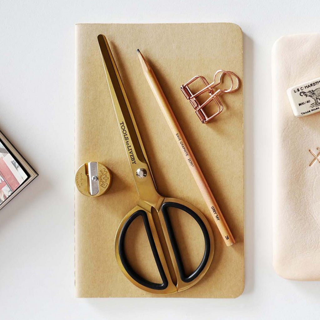 A styled flat-lay of stationery items on a light surface: a gold-handled scissors with black handles, a matching gold pencil, a rose-gold binder clip, a small gold sharpener, and a beige notebook.