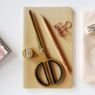 A styled flat-lay of stationery items on a light surface: a gold-handled scissors with black handles, a matching gold pencil, a rose-gold binder clip, a small gold sharpener, and a beige notebook.