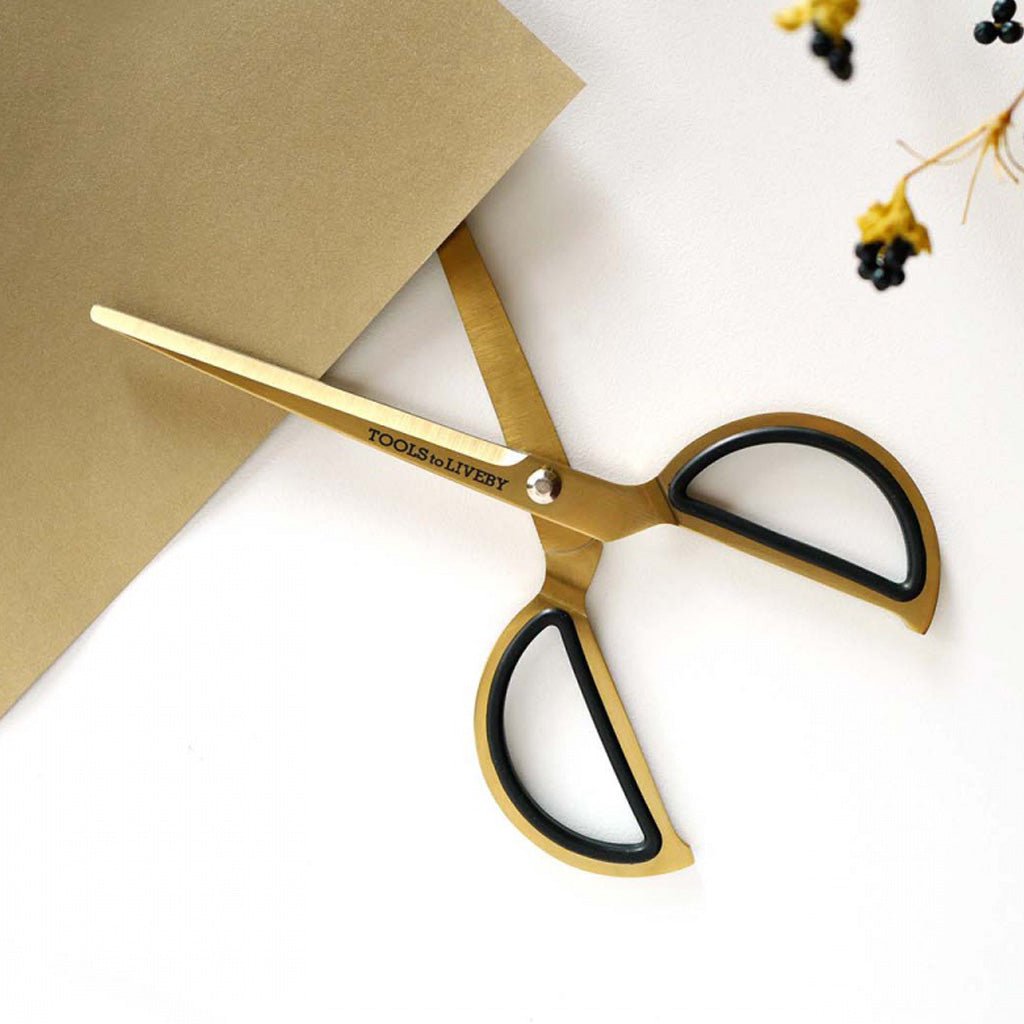 Gold-toned scissors with black inner handles and the brand name 'TOOLS OF LIVEBY' printed on the blade, styled on a white surface with a brown paper corner and small dried flowers.
