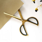 Gold-toned scissors with black inner handles and the brand name 'TOOLS OF LIVEBY' printed on the blade, styled on a white surface with a brown paper corner and small dried flowers.