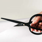 A close-up of a hand holding a pair of black scissors with the brand name 'TOOLS to LIVEBY' printed on the blade, cutting a sheet of white paper against a light neutral background.