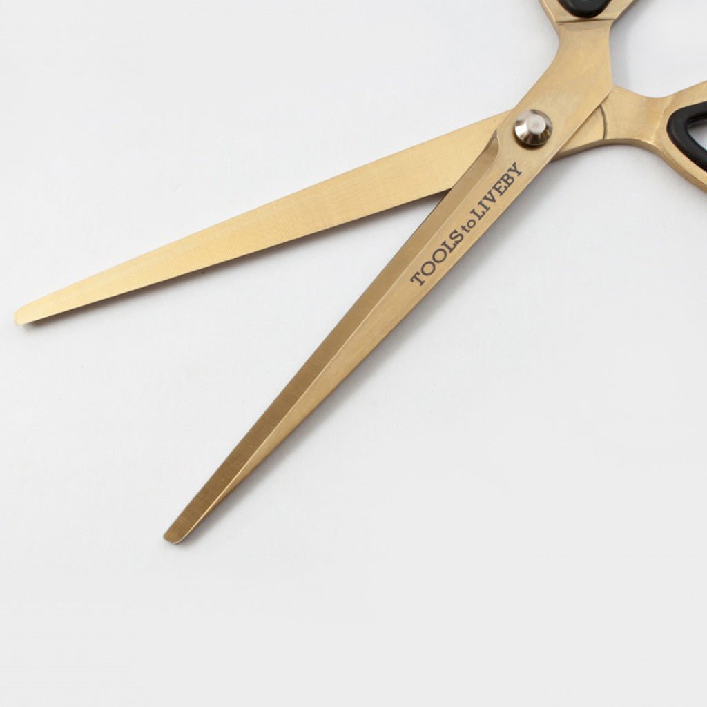 Gold-colored scissors with black handles and a silver pivot screw, featuring the text 'TOOLS TO LIVEBY' printed on one blade, set against a plain light gray background.