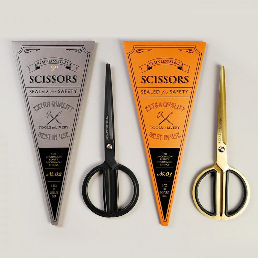 Gold-toned scissors with black rubberized finger loops and matte black scissors with rounded handles next to packaging on gray background