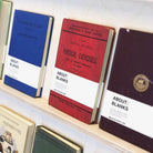 A close-up of vintage books displayed on a wooden shelf, featuring blue, red, and dark purple covers with white bands labeled 'ABOUT: BLANKS'.