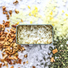 Open tin of aegean salt blend surrounded by spices on white background
