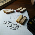 Wooden monogram rubber stamps with engraved letters K and M, shown alongside a black ink pad on a graph paper surface with a coffee-themed note.