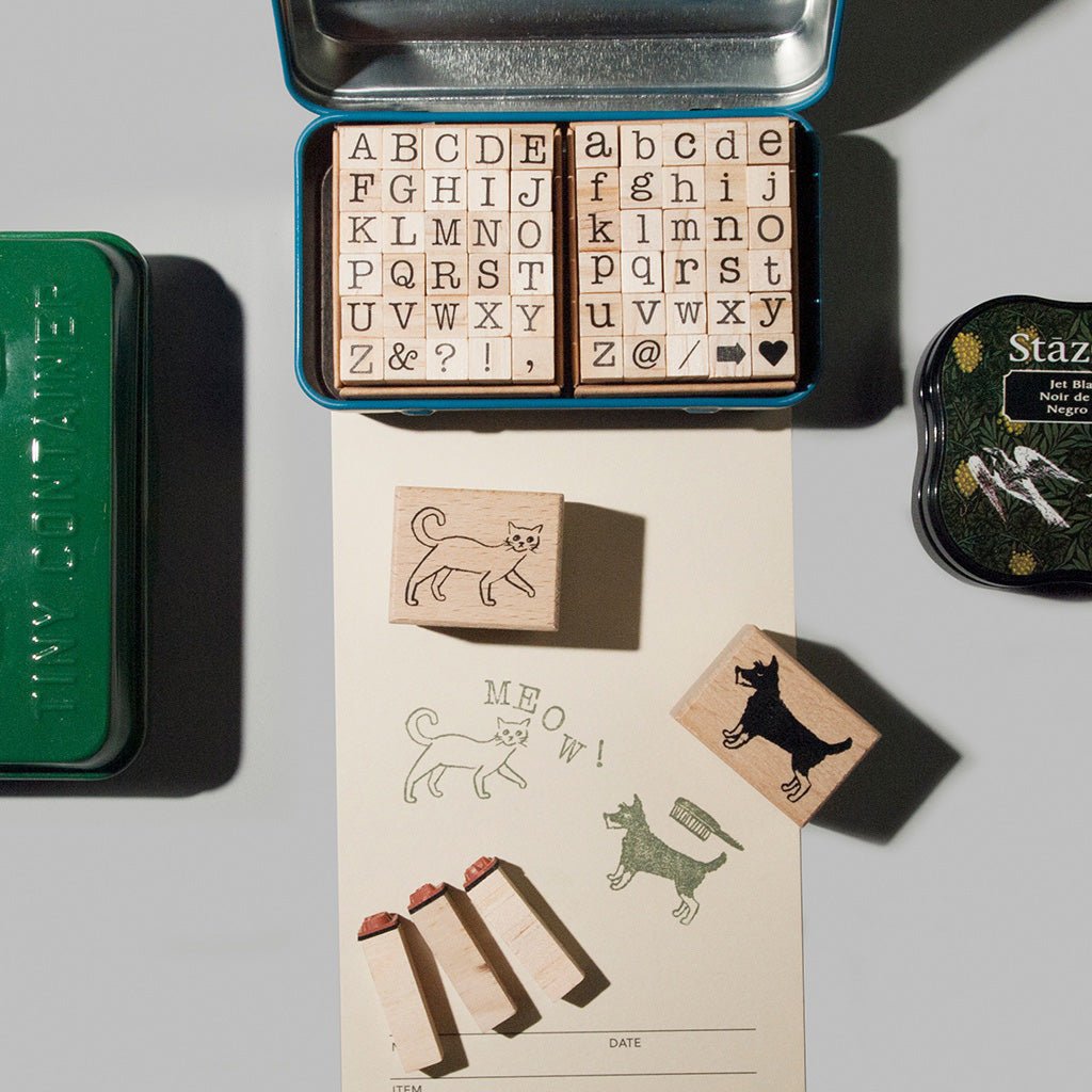 A set of wooden rubber stamp blocks in a blue metal tin, shown with several stamped animal and letter examples on a sheet below, alongside a green tin and a round stamp ink pad on a light gray background.