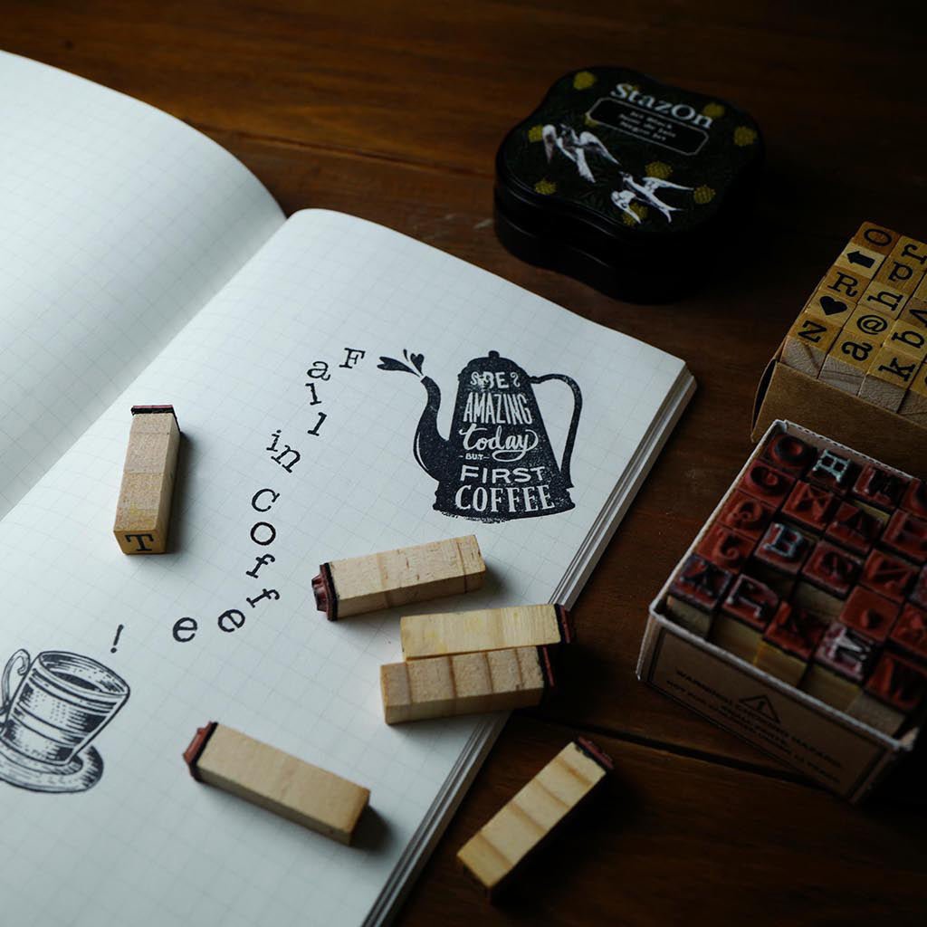A styled flat-lay of wooden rubber stamp blocks arranged around an open notebook on a dark wooden table. The notebook shows a stamped coffee-themed illustration and text.