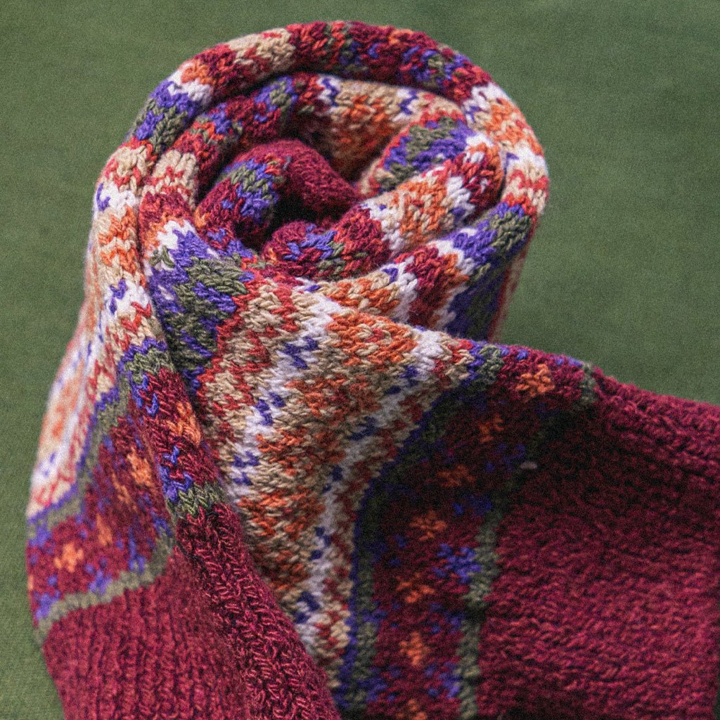 A close-up of a knitted fair isle sock with red, orange, purple, white, and green knit details against a green background.
