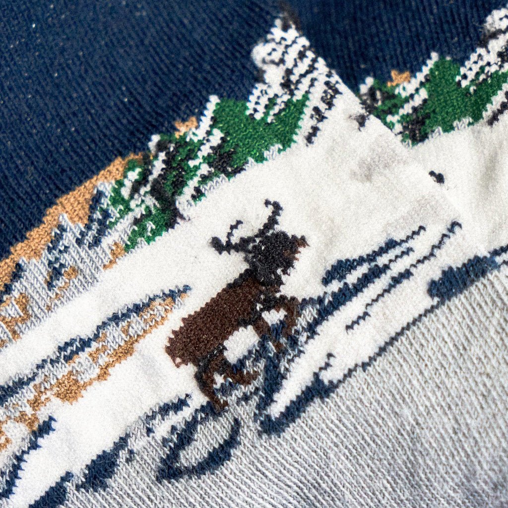 Close-up of a knit sock showing a winter landscape pattern with a brown reindeer on a snowy white background.
